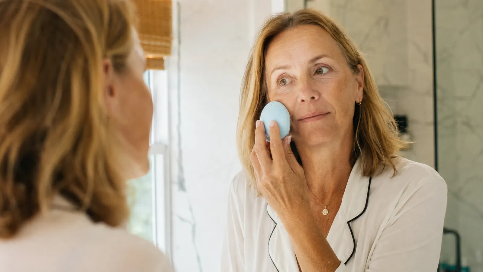 Woman in her 50s gently using a silicone facial cleansing brush in a bathroom, soft morning light, relaxed expression