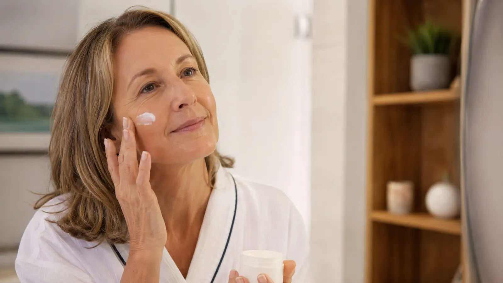 Woman in her 50s applying gel cream to her face in front of a bathroom mirror, soft morning light
