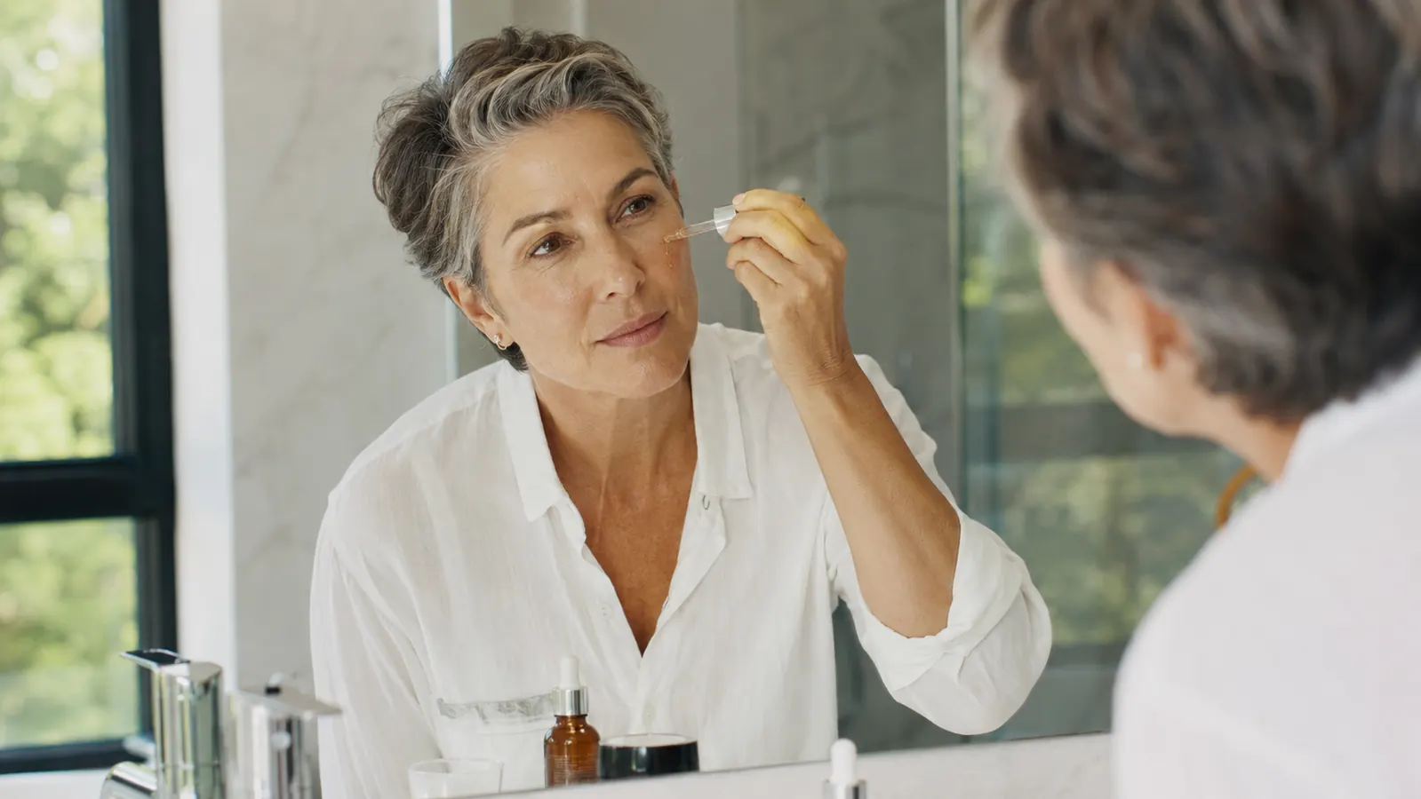 Woman in her fifties applying serum in a bright, clean bathroom mirror